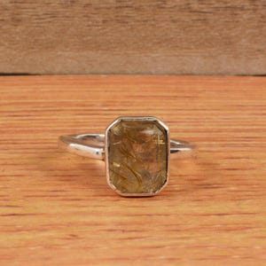 Pode incluir: Um anel de prata com uma pedra preciosa retangular castanho-dourada. A pedra preciosa tem inclusões douradas internas. O anel está sobre um fundo de madeira.