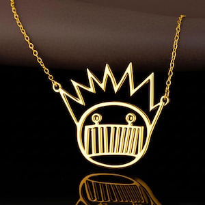 Puede incluir: Un collar dorado con un colgante que representa una cara estilizada con una corona. El colgante tiene una cara circular con líneas verticales para la boca y dos pequeños círculos para los ojos. La cadena es delicada y dorada.