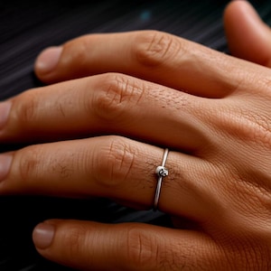 Könnte beinhalten: Ein silberner Ring mit einem kleinen, runden Akzent, möglicherweise ein Edelstein. Der Ring ist dünn und wird an einem Finger getragen. Das Bild konzentriert sich auf den Ring.