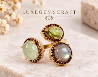 May include: A gold-toned open ring featuring three distinct cabochon gemstones: a round pale green stone, a pear-shaped light green stone, and a round grey iridescent stone. Each stone is surrounded by a halo of small dark accent stones.