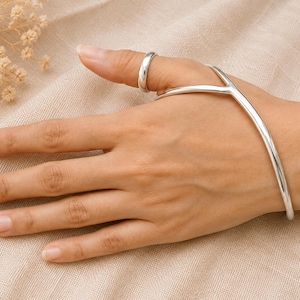 May include: A polished silver-tone hand ornament consisting of a smooth, plain band ring connected by a slender metal bar to a curved, open-ended bracelet that encircles the wrist. The piece features a sleek, continuous metallic finish.