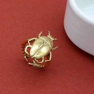 May include: A gold-colored ring in the shape of a beetle, with intricate details on the body and legs. The ring is set against a red background, with a white ceramic object in the background.