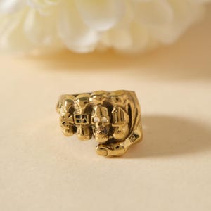 May include: A gold-colored ring featuring a clenched fist design with skull details. The ring has a detailed texture and is set against a soft, neutral background. The ring's design incorporates skull motifs, adding a unique and edgy aesthetic.
