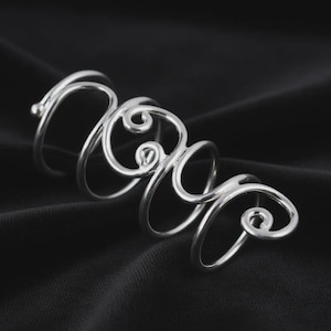 May include: A silver-colored ring with a unique, swirling design. The ring features multiple loops and curls, creating an abstract, artistic look. The jewelry is displayed against a dark background.