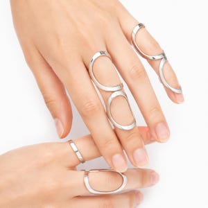 May include: A set of silver-toned metal rings featuring open, elongated oval designs. These rings are worn on multiple fingers, with some encircling the base of the finger and others extending along the length of the finger with a smooth finish.