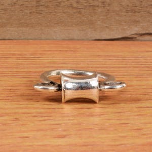 Könnte beinhalten: Ein silberfarbener Metallring mit einem zylindrischen Mittelteil und zwei kleinen, abgerundeten Verlängerungen. Der Ring ist vor einem warmen Holzgrund platziert, der seinen metallischen Glanz und sein schlichtes Design hervorhebt.