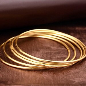 Può includere: Un set di cinque braccialetti circolari sottili color oro impilati insieme. Ogni braccialetto presenta una superficie metallica liscia e lucida con una finitura riflettente. I braccialetti sono disposti in una formazione sovrapposta.