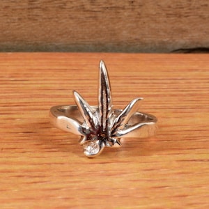 Puede incluir: Un anillo de plata con un diseño de hoja de cannabis. El anillo tiene un acabado pulido y se encuentra sobre una superficie de madera. La hoja es el elemento central, con vetas detalladas y una apariencia tridimensional.