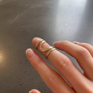 May include: A gold-colored finger ring with a unique, flowing design. The ring wraps around the finger with a series of curved lines, creating an elegant and modern look. The ring is made of a shiny, metallic material.