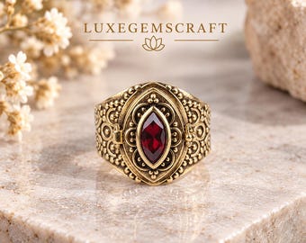 May include: A gold-toned metal ring featuring an intricate, embossed filigree pattern and a central marquise-cut deep red gemstone. The band displays a textured, ornate design with small beaded details surrounding the central stone setting.
