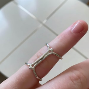 May include: A silver-colored ring with a unique design. The ring features a rectangular center with curved, upward-pointing arms on each end, and small spherical accents. The ring is worn on a finger.