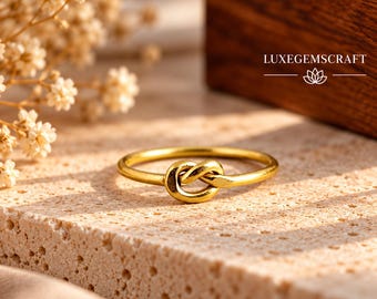 May include: A gold-toned ring featuring a central knot design. The band is smooth and circular, with the knot element positioned in the center. The text LUXEGEMSCRAFT is visible in the upper right corner, accompanied by a small lotus flower icon.