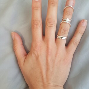 May include: A hand with several silver rings on the fingers. The rings include a simple band, a ring with a square stone, and a curved ring. The hand is resting on a light gray surface.