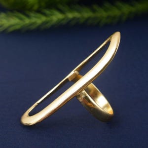 May include: A gold-colored ring with a unique design. The ring features a band and an elongated, open oval shape that extends upwards. The ring is set against a dark blue background, highlighting its metallic sheen.