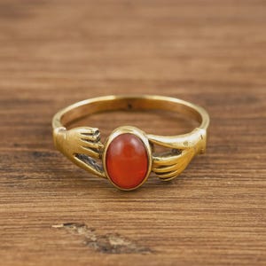 May include: A gold-colored ring featuring a central oval, reddish-orange stone. The ring's band is designed with two stylized hands on either side of the stone, creating a unique and artistic look. The ring is set against a wooden background.