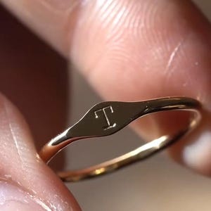 Könnte beinhalten: Ein goldfarbener Metallring mit einer kleinen, flachen, ovalen Oberfläche, in die der Großbuchstabe T eingraviert ist. Das Band ist dünn und poliert, wobei der Text "wear their initial on your pinky" über dem Ring im Bild zu sehen ist.