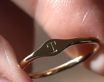 Könnte beinhalten: Ein goldfarbener Metallring mit einer kleinen, flachen, ovalen Oberfläche, in die der Großbuchstabe T eingraviert ist. Das Band ist dünn und poliert, wobei der Text "wear their initial on your pinky" über dem Ring im Bild zu sehen ist.