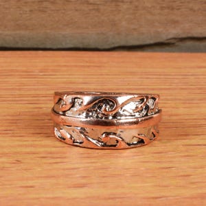 May include: A rose gold ring with a detailed wave pattern. The ring is set against a wooden background, showcasing its intricate design. The band features a continuous, flowing wave motif, adding a touch of elegance.