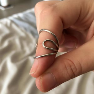 Könnte beinhalten: Ein silberfarbener Ring mit einem einzigartigen, fließenden Design. Der Ring umschließt den Finger mit geschwungenen und geschlungenen Elementen und erzeugt eine abstrakte, moderne Ästhetik. Der Ring besteht aus glänzendem Metall.