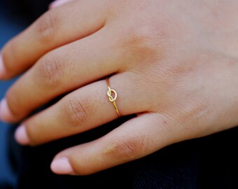 May include: A delicate gold ring with a knot design. The ring is thin and sits on a finger, showcasing its simple yet elegant style. The ring's warm gold colour contrasts with the skin tone, highlighting the jewellery's design.