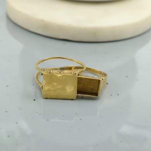 Op de afbeelding: Close-up van drie goudkleurige ringen. Twee ringen hebben een vierkante zetting en één heeft een rechthoekige zetting. De ringen liggen op een lichtgrijze ondergrond. De ringen zijn waarschijnlijk voor het maken van sieraden of knutselen.