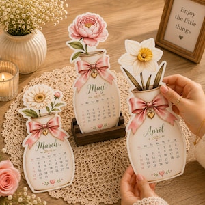 Puede incluir: Tres calendarios de papel troquelado para marzo, abril y mayo de 2026, cada uno con forma de jarrón con un arreglo floral, un lazo rosa y un dije de corazón. El calendario de marzo muestra una margarita, abril un narciso y mayo una peonía.