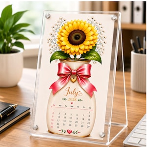 May include: A clear acrylic desktop calendar frame displaying a July 2028 page. The design features a large yellow sunflower with white baby's breath in a cream-coloured vase, accented by a pink ribbon bow and a small gold heart charm at the centre.