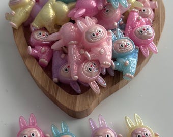 May include: A collection of colorful, cartoonish bunny figurines in various pastel shades, including pink, blue, yellow, and purple. The bunnies are inside a wooden heart-shaped dish, with some arranged outside. The figurines have a smiling face.