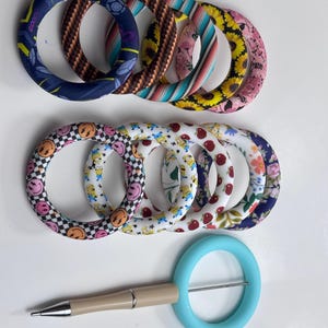May include: A selection of patterned wristlet keyrings and a light blue pen with a beige barrel. The keyrings display diverse designs, including floral, smiley faces, and geometric patterns. The pen has a silver tip and a light blue ring.