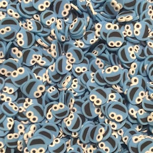 Peut inclure: Une grande collection de petites décorations en sucre bleues, plates et circulaires. Chaque pièce présente un motif imprimé de visage souriant avec deux yeux blancs et une bouche noire, rappelant un personnage monstre bleu pour pâtisseries.