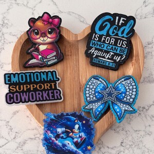 Peut inclure: Un plateau en bois en forme de cœur présente cinq épingles colorées. Les épingles représentent un écureuil de dessin animé avec des lunettes, un nœud bleu avec une étoile, le texte "Emotional Support Coworker", un graphique Sonic et le texte "If God is for us, who can be against us?"