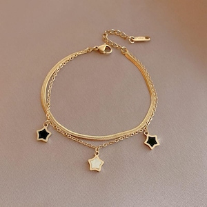 Peut inclure: Bracelet doré à deux chaînes délicates. Le bracelet comporte trois breloques en forme d'étoile, deux noires et une blanche, suspendues aux chaînes. Le bracelet est présenté sur une surface de couleur neutre.