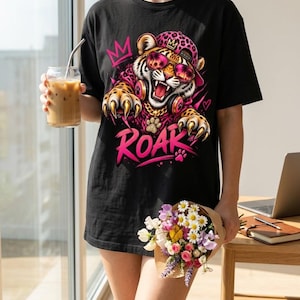 Könnte beinhalten: Ein schwarzes T-Shirt mit einer Grafik eines Tigers, der eine pinke Kappe mit Leopardenmuster, eine Sonnenbrille und Kopfhörer trägt. Der Tiger hat seine Krallen ausgefahren und das Wort ROAR ist in fetten pinken Buchstaben unter dem Tiger gedruckt.