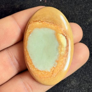 May include: An oval gemstone with a polished surface. The stone has a central pale green area, encircled by a band of orange and tan shades. The outer edge is a light tan colour. The gemstone is held in a hand.