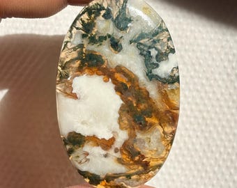 May include: Oval-shaped gemstone with a unique pattern. The stone features a mix of white, brown, green, and yellow hues, creating a natural, artistic design. The gemstone is held between fingers, showcasing its size and shape.