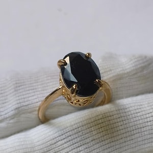 May include: A gold-colored ring featuring a large, oval-cut black gemstone. The ring has a delicate, filigree design on the band near the gemstone setting. The gemstone is the focal point, reflecting light and adding a touch of elegance to the jewelry.