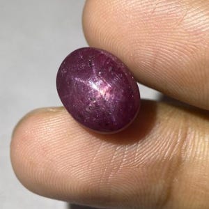 May include: A polished, oval ruby gemstone. The gemstone is a deep, purplish-red colour with a star-like effect visible on its surface. The star effect is created by light reflecting off of inclusions within the stone. The ruby is held between two fingers.