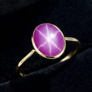 Può includere: Un anello d'oro con una grande gemma di rubino a stella rosa-viola a taglio ovale. Il rubino a stella presenta un motivo a stella a sei raggi. L'anello è incastonato in una semplice ed elegante fascia d'oro, esposto in un portagioie nero.