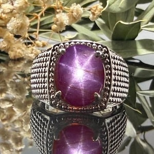 Könnte beinhalten: Ein Silberring mit einem großen, ovalen, tief magentafarbenen Sternsaphir-Cabochon in der Mitte. Der Stein zeigt ein deutliches sechsstrahliges Sternmuster. Die Ringschiene hat eine texturierte, kreuzschraffierte Metalloberfläche mit Perlenrand.