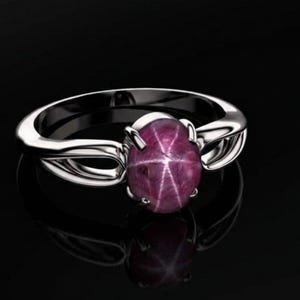 Op de afbeelding: Een zilveren ring met een grote, ovale ster-robijn edelsteen. De robijn heeft een dieproze kleur met een zesstralig sterpatroon. De ring heeft een split-schacht ontwerp.