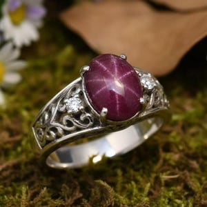 Può includere: Un anello d'argento con una grande gemma ovale rosso scuro che mostra un effetto a stella. L'anello è adornato con piccole pietre di accento trasparenti e dettagli di filigrana intricati. L'anello è posto su muschio verde e fiori.