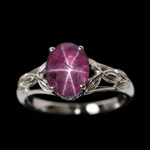 May include: A silver ring with a large, oval, purplish-red gemstone displaying a star effect. The ring's band has an intricate, openwork design. The gemstone is the central feature, set against a black backdrop.