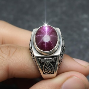 May include: A silver ring featuring a large, oval-shaped, deep red gemstone with a star-like pattern. The ring has intricate side detailing and a decorative front design. The gemstone is the focal point, set in a polished silver band.
