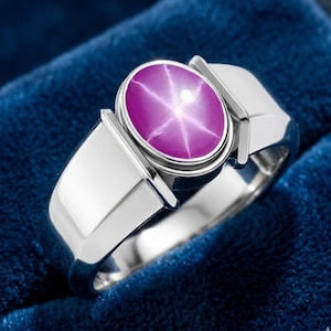 May include: A silver ring featuring a large, oval-cut, purplish-pink star sapphire gemstone. The gemstone has a six-ray star pattern. The ring has a polished, modern design with a wide band and rectangular shoulders.