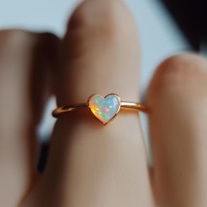 Peut inclure: Une bague dorée ornée d'une opale en forme de cœur. L'opale présente un jeu de couleurs vibrant, avec des éclats de vert, de bleu et d'orange. Le corps de la bague est fin et délicat, créant un design minimaliste. La bague est un bijou.
