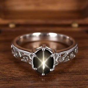 Puede incluir: Un anillo plateado con una gema ovalada con efecto de estrella. El anillo tiene un diseño calado ornamental en la banda. La gema es de color verde oliva oscuro. El anillo se muestra sobre un fondo de madera.