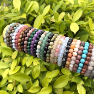 May include: A collection of beaded bracelets in various colors, including white, red, brown, green, purple, black, yellow, orange, blue, and gray. The bracelets are made of small, round beads and are displayed against a backdrop of green foliage.