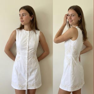 May include: A sleeveless white mini dress featuring a front button-down closure and a round neckline. The dress is adorned with delicate beige floral embroidery along the side panels and the bottom hemline, creating a structured silhouette.
