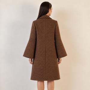 May include: A brown coat featuring a small-scale houndstooth pattern, a structured collar, and wide, flared bell sleeves. The garment is knee-length and displays a clean, tailored silhouette with a vertical seam running down the center back.
