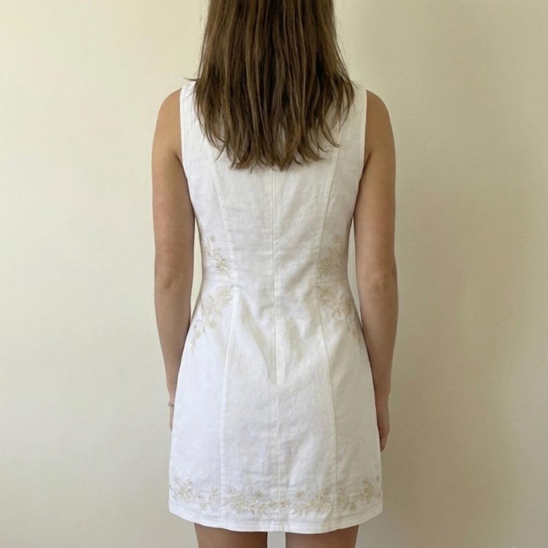 May include: A sleeveless white mini dress featuring a fitted bodice and a straight hemline. The garment is decorated with subtle, light-coloured floral embroidery patterns along the side panels and the lower hem area, creating a cohesive design.